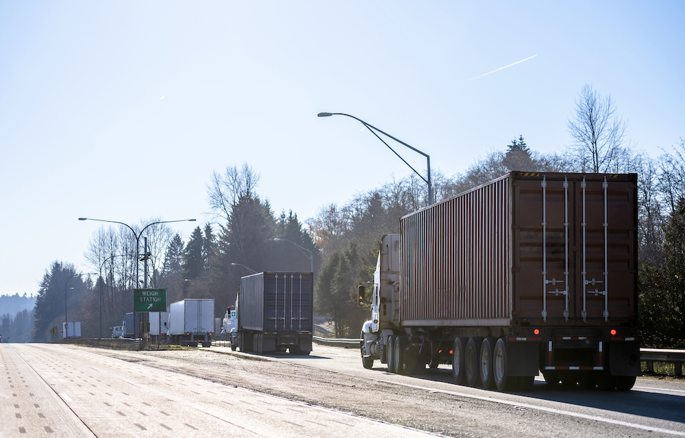 Different big rigs, semi trucks with containers, and semi trailers standing in line to weight station for check truck and cargo weight and axle weight distribution next to highway.